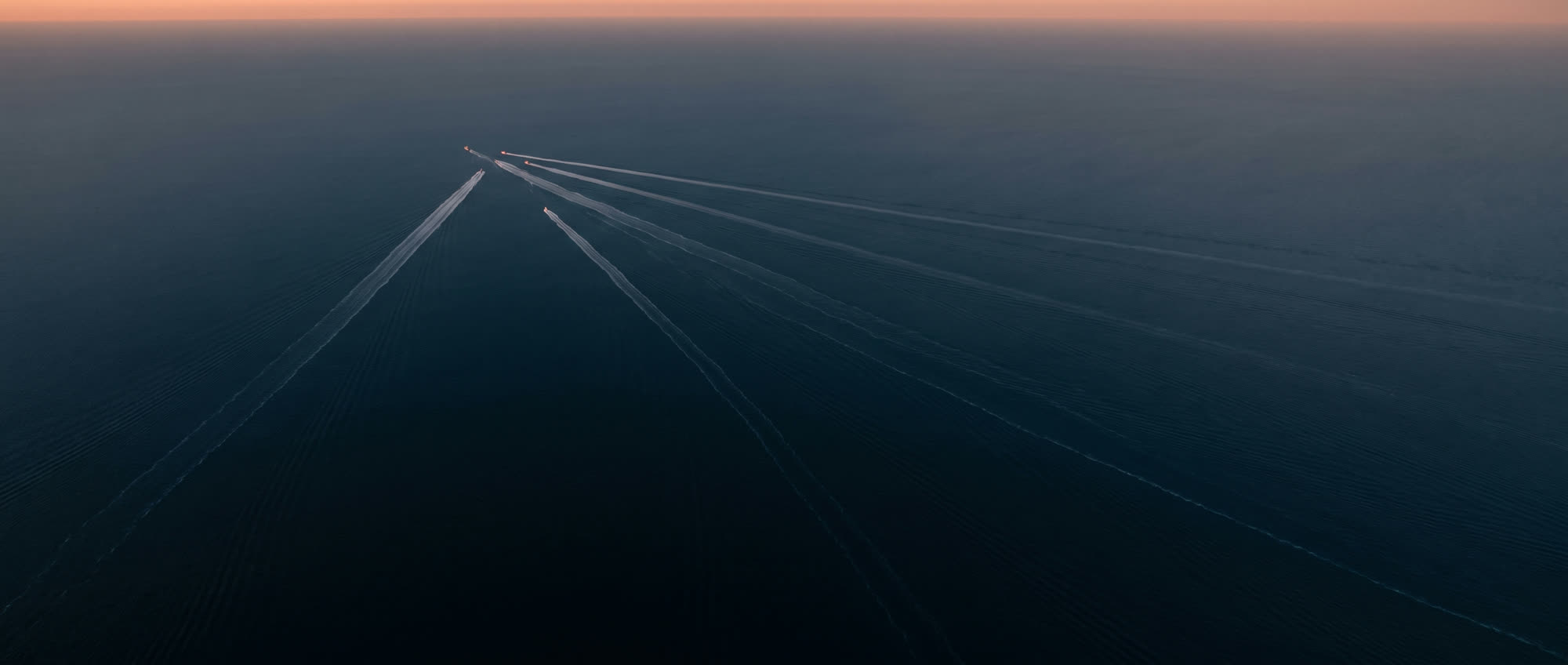 Dark open sea from high altitude at first light, several long wakes converging on a single point