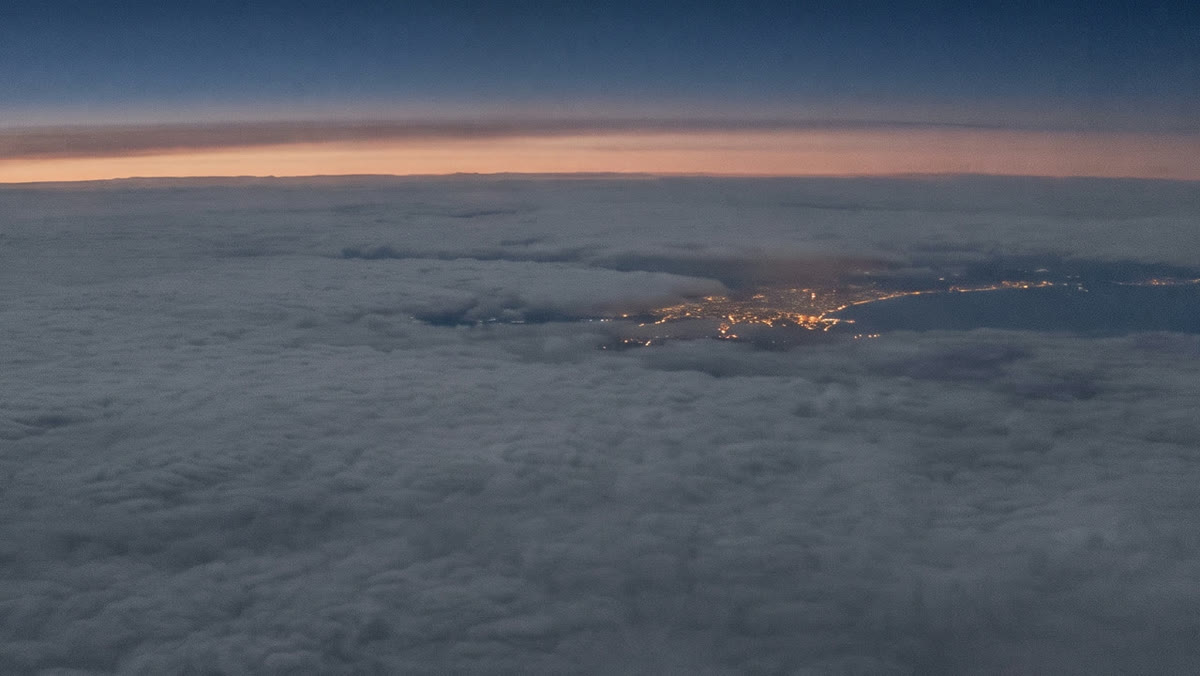 City lights glowing through a gap in the clouds below, with the dawn horizon above, seen from high altitude.
