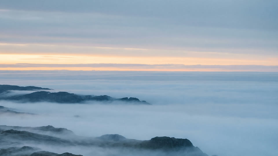 Low fog filling the valleys between dark hills, under a pale dawn sky.