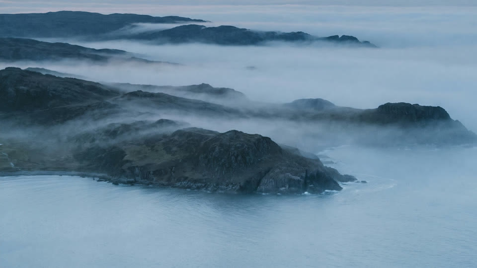 Forested headlands rising out of fog over calm water.
