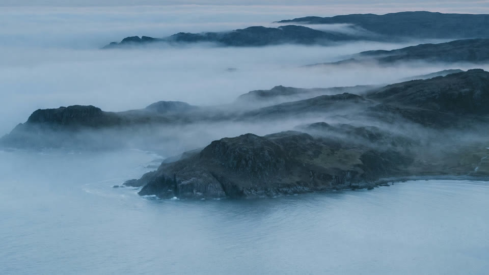 Forested headlands rising out of fog over calm water.