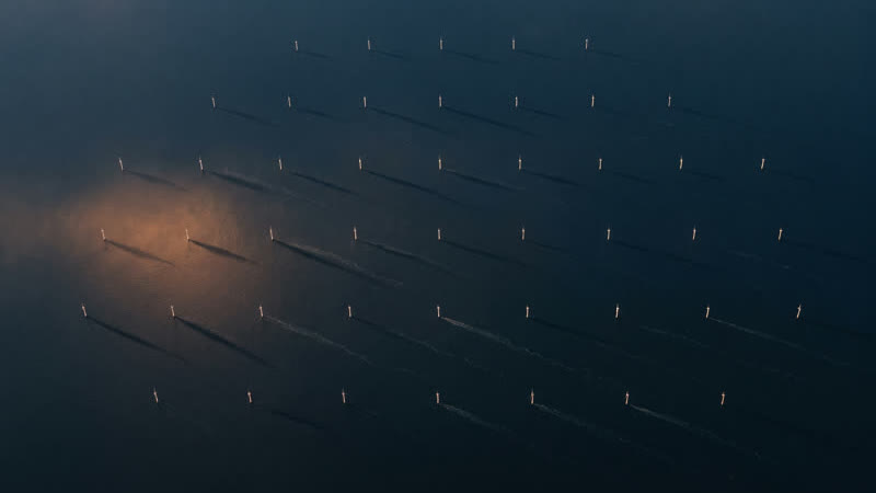 Rows of wind turbines at dusk with a warm glow in the distance, seen from above.
